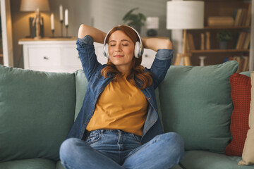 Obraz premium A young woman sits back on a comfortable couch, wearing headphones, and appears content as she listens to music. The cozy living room has soft lighting and warm decor.