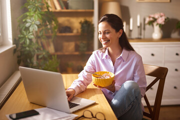A woman smiles as she eats a bowl of cereal while working on her laptop in a well-lit home office....