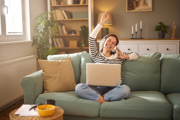 Woman sitting cross-legged on a green couch, smiling joyfully while using a laptop in a calm and...