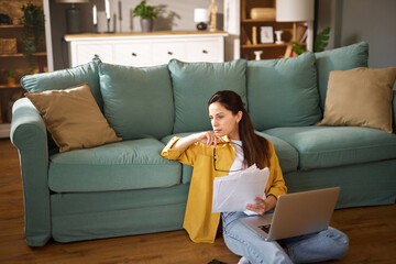 A young woman sits comfortably on a couch in her home office, reviewing documents while holding...