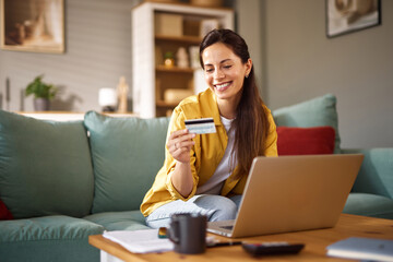 A woman sits comfortably on a couch, using a laptop while holding a credit card, smiling as she...