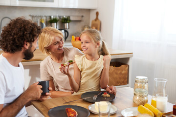 A joyful family shares a light meal at home, with parents engaging with their cheerful daughter who...
