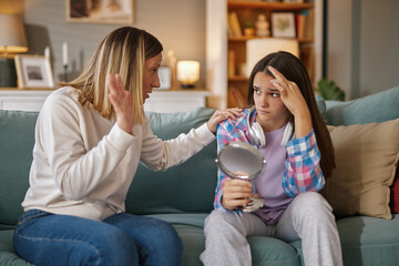 A teenage girl sits on a couch looking troubled while holding a mirror, as her mother gestures...