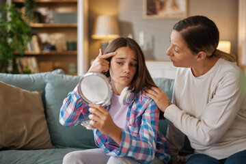 A teenage girl examines her reflection in a mirror with a worried expression, while her mother...