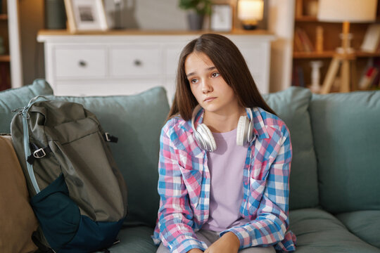 A teenager sits on a couch looking pensive and withdrawn. She is wearing a plaid shirt with headphones around her neck, a backpack beside her, reflecting feelings of isolation. - Powered by Adobe