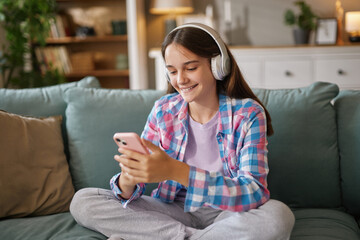 Obraz premium A happy teenage girl with headphones smiles as she listens to music on her smartphone while sitting cross-legged on a soft couch in a bright living room.