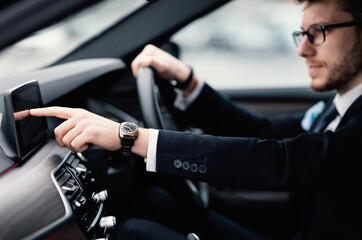 Adult businessman in formalwear and eyeglasses driving car and touching dashboard with finger,...