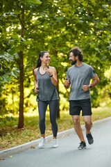 A man and woman jog side by side on a sunny day, surrounded by lush greenery, engaging in...