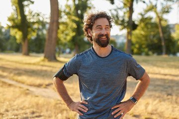 A man stands smiling in a park, hands on his hips, after a refreshing jog. The golden sunlight...