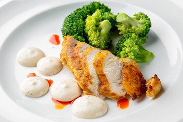 Grilled chicken breast with steamed broccoli and white sauce, garnished with red hot sauce on a white plate. Healthy and appetizing meal presentation.