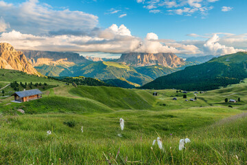 Panoramic view of green meadow and cabins with Sassolungo mountains under dramatic clouds in Italian Dolomites Alps, Trentino Alto Adige, Italy. Alpine meadow of Seiser Alm with Langkofel