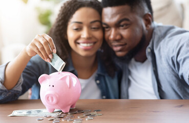 Family Budget. Happy Black Couple Putting Money Cash To Piggybank, Making Savings For Future, Sitting Together At Home