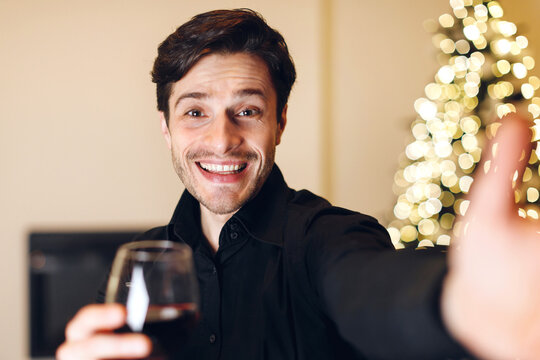 Cheers. Portrait of cheerful happy young bearded guy holding glass, drinking red wine, toasting and talking selfie, making video call, reaching hand to camera, celebrating at party, blurred background - Powered by Adobe