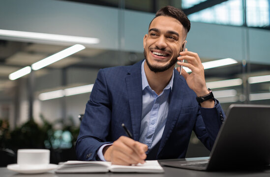 Positive young arab guy in suit businessman working at modern office, sitting at worktable in front of laptop, talking on mobile phone and taking notes, looking at copy space and smiling - Powered by Adobe