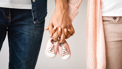 Closeup of pregnant couple carrying small baby shoes while holding hands together, unrecognizable...