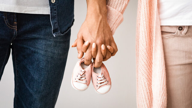 Closeup of pregnant couple carrying small baby shoes while holding hands together, unrecognizable loving man and woman expecting baby, enjoying future parenthood, cropped image, free space - Powered by Adobe