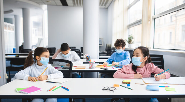 Multicultural schoolchildren wearing disposable medical masks sitting at table in classroom, keeping new normal social distance, looking at each other, studying at elementary school and writing - Powered by Adobe