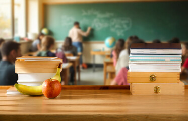 School desk with school supplies and fruit lunch, blurred students and teacher in classroom background. School-related atmosphere of learning, back-to-school excitement and education in progress.