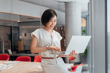 Smiling mature Asian professional it specialist businesswoman working on laptop pc standing in office workplace. 30s middle-aged woman using computer technology app for banker financial work online.