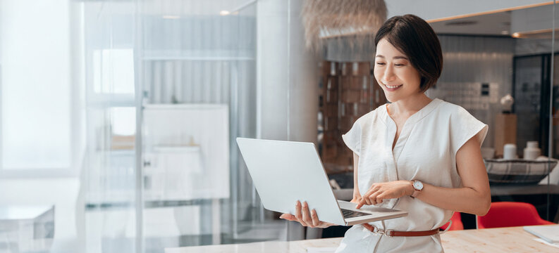 Asian professional it specialist businesswoman working on laptop pc standing in office workplace. Middle-aged woman using computer technology app for banker financial work online. Banner, copy space
