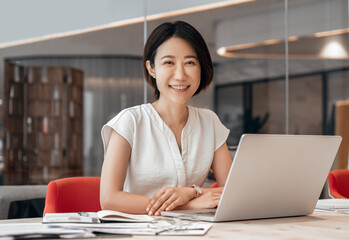 Smiling portrait at camera mature Asian professional businesswoman ceo analyst working on laptop pc at desk in office. Middle-aged woman using computer technology for financial marketing work online.