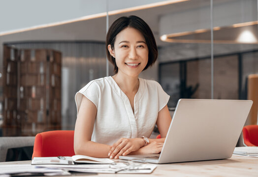 Smiling portrait at camera mature Asian professional businesswoman ceo analyst working on laptop pc at desk in office. Middle-aged woman using computer technology for financial marketing work online.