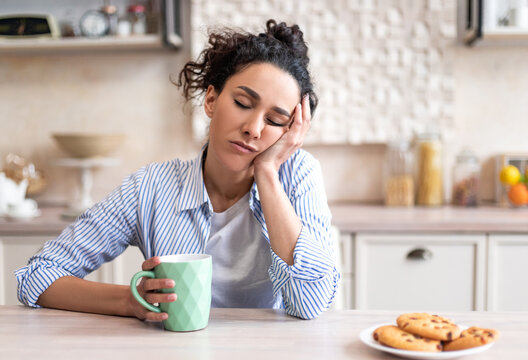 Sleepy young woman sitting at dining table in kitchen with closed eyes, holding cup while having breakfast. Early wake up, insomnia, lack of sleep