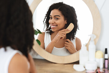 Attractive African Woman Combing Her Curly Hair In Front Of Mirror At Home, Smiling And Enjoying...