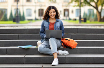 Joyful young indian woman student studying online, using laptop computer outdoors, sitting on stairs at college campus, doing homework, copy space. E-learning concept