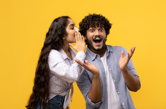 Young indian woman sharing secrets with her excited boyfriend, millennial couple whispering gossips to each other over yellow studio background