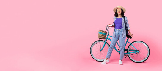 Springtime Activity. Full body length shot of trendy asian lady wearing summer hat and backpack posing and holding handlebar of classic bicycle with wicker basket isolated on pink studio background