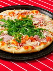 A pizza with ham and spinach on a black tray. The pizza is sitting on a red and white stripe table cloth. Fine Italian cuisine with fresh ingredients ready to be cooked. Fast food meal.