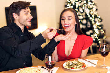 Romantic Date Concept. Smiling handsome guy feeding his cute girlfriend with pasta. Portrait of happy excited family sitting at table and eating meal at restaurant or at home, drinking red wine