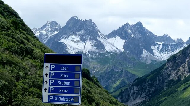 Snow-capped alpine peaks rise majestically above green valleys, with a roadside parking sign pointing toward Lech, Z&uuml;rs, and Stuben in the Austrian Alps.