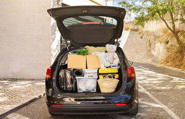 Moving boxes and suitcases, various belongings, plants in open trunk of black car outdoor on the street next to building, no people, copy space. Relocation, moving, real estate, new beginning concept © Prostock-studio