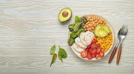 Healthy bowl with grilled chicken breast, avocado, chickpeas, corn, tomatoes and mixed greens, top view on a white wooden table from above, a balanced and nutritious meal