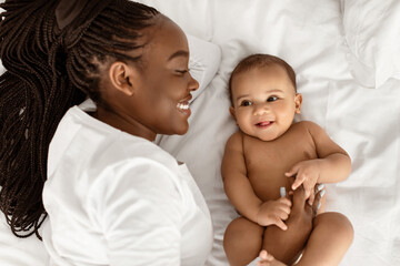 Above top high angle view portrait of smiling black young mother with long afro braids playing with her adorable small baby, lying on bed, touching infant's belly and spending time with kid in bedroom