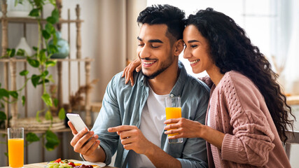 Cheerful Arab Spouses Using Smartphone While Eating Breakfast In Kitchen, Happy Middle Eastern Couple Enjoying Tasty Morning Meal, Shopping Online Or Checking Social Networks On Cellphone, Free Space