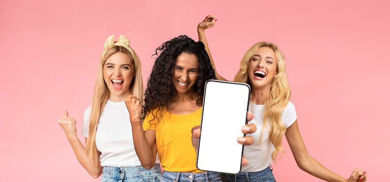 Carefree attractive millennial ladies in stylish casual outfits having fun together on pink studio background, showing smartphone with empty screen, dancing and laughing, mockup, panorama - Powered by Adobe