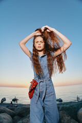young woman in denim jumpsuit stands by the sea at sunset with long hair styled confidently and a...