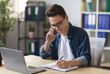 Young Office Manager Talking On Mobile Phone And Taking Notes To Notepad, Millennial Male Employee...