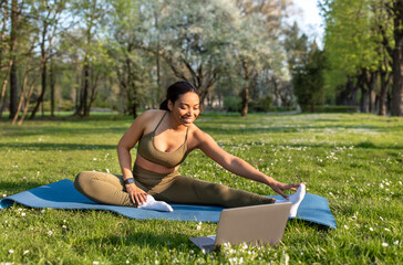 Sporty young black lady stretching her leg near laptop pc, following online sports video tutorial...