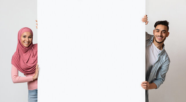 Smiling young muslim couple holding white advertisement board, demonstrating free copy space for your text or design, positive arab man and woman in hijab standing over grey background, panorama - Powered by Adobe