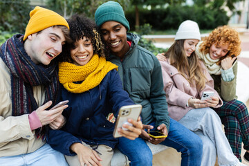 Young group of teenage people using smartphones together at city street on winter. Millennial student friends relaxing outside browsing on internet and taking selfies with phones. Youth concept.