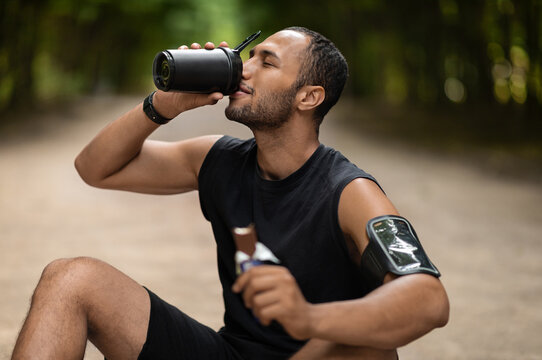 Tired middle eastern young guy sportsman sitting on the ground at public park, having snack while training outdoors, drinking protein cocktail and eating protein bar, copy space. Sports nutrition - Powered by Adobe