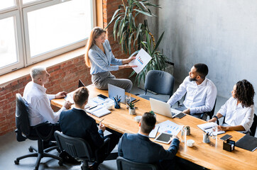 Corporate Meeting. Business Lady Showing Charts To Coworkers Analyzing Company Growth Sitting On Table In Modern Office