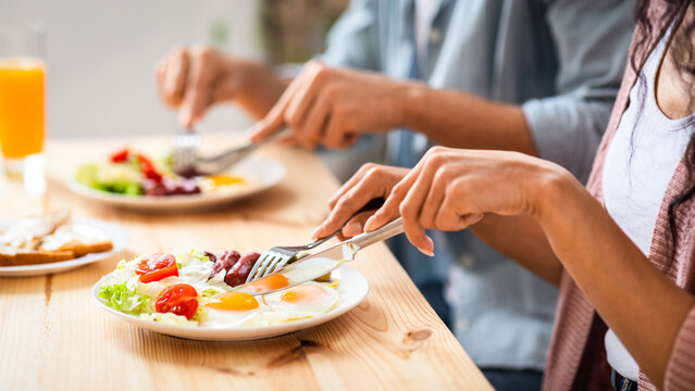 Unrecognizable couple using fork and knife while eating tasty breakfast in kitchen, cropped image of man and woman enjoying morning meal, having plate with fresh salad, eggs and sausages, closeup