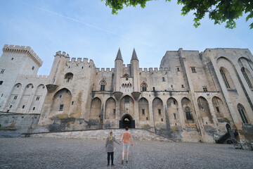 Married Couple holding hands facing the grand Gothic facade of the Papal Palace in Avignon, symbol of medieval history, political change and chistanitiy and papacy.