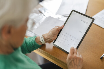 Senior woman using calculator app on digital tablet at table 
