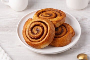 Cinnamon bun in white plate with cinnamon sticks, coffee and napkin on white table